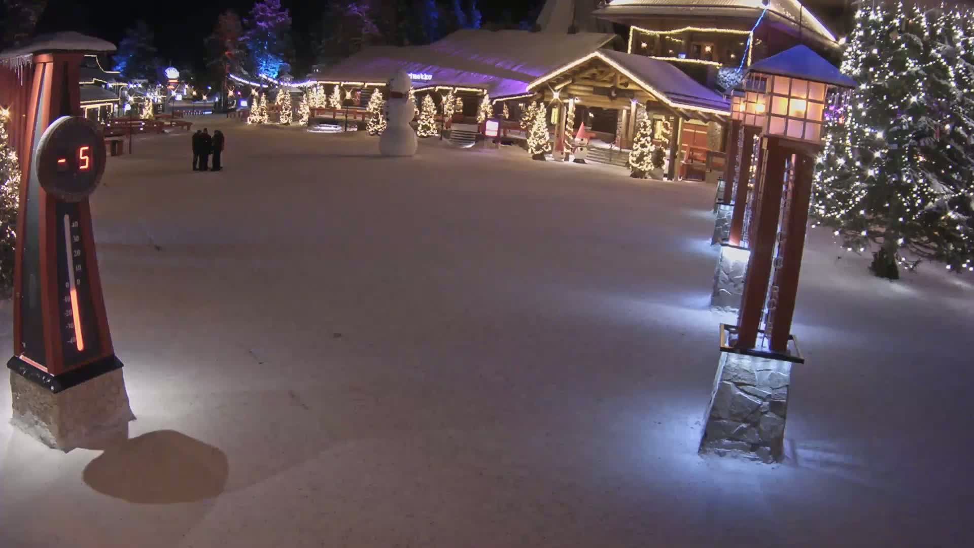 A snow-covered village square at night, illuminated by lights on buildings and lampposts, shows a thermometer reading -5 degrees, a snowman, and several people.