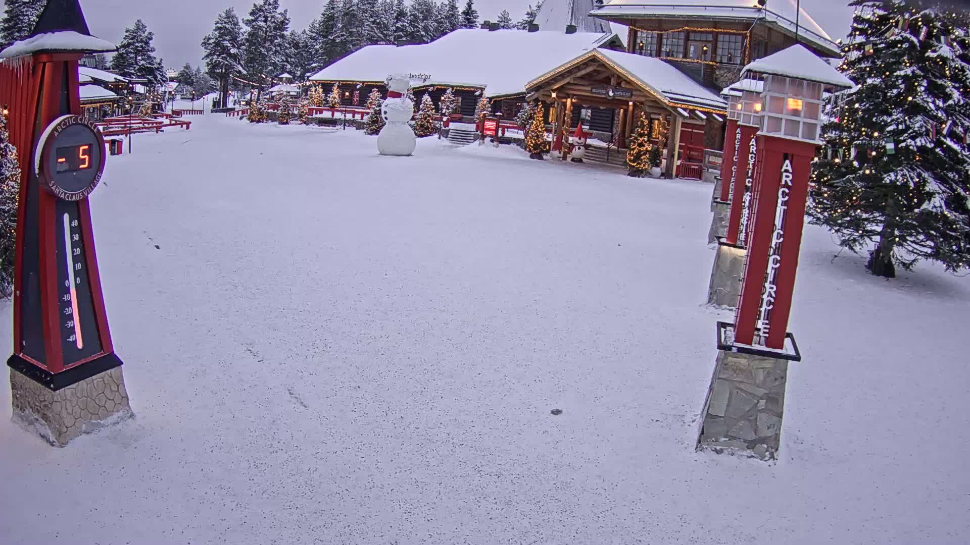 A snow-covered plaza with a large snowman, Christmas trees, and buildings, under an overcast sky, shows a temperature of -5 degrees.