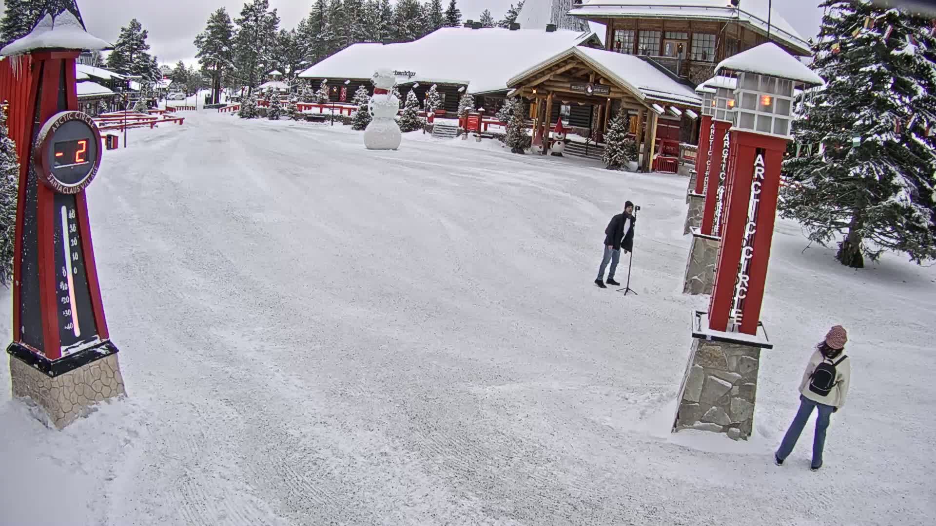 A snow-covered plaza features two people, a large snowman, several tall red and white signs, and a building in the background, with a temperature of -2 degrees displayed on a thermometer.