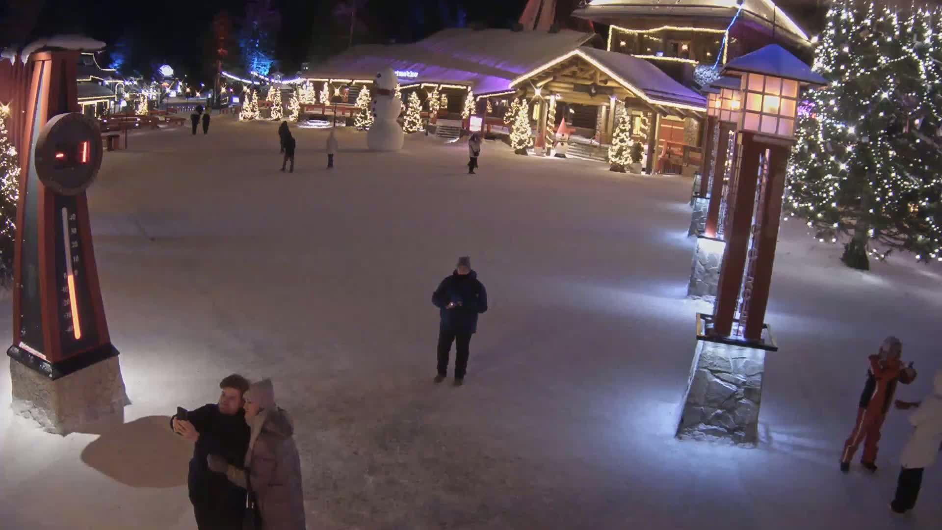A snow-covered village square at night, illuminated by lights on buildings and lampposts, features several people walking and a couple taking a selfie near a large thermometer showing a temperature below zero.