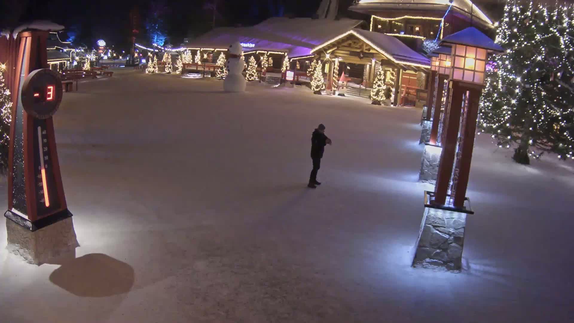 A snow-covered plaza at night, illuminated by lights on buildings and lampposts, features a person standing alone near a large thermometer that reads -3 degrees, and a snowman stands near a festively decorated building in the background.