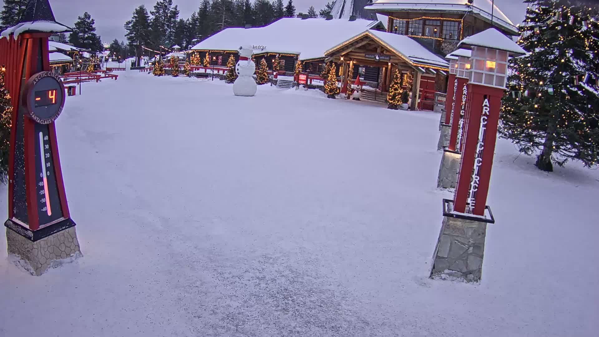 A snow-covered plaza with a wooden building decorated with Christmas lights, a snowman, and a thermometer showing a temperature of -4 degrees.