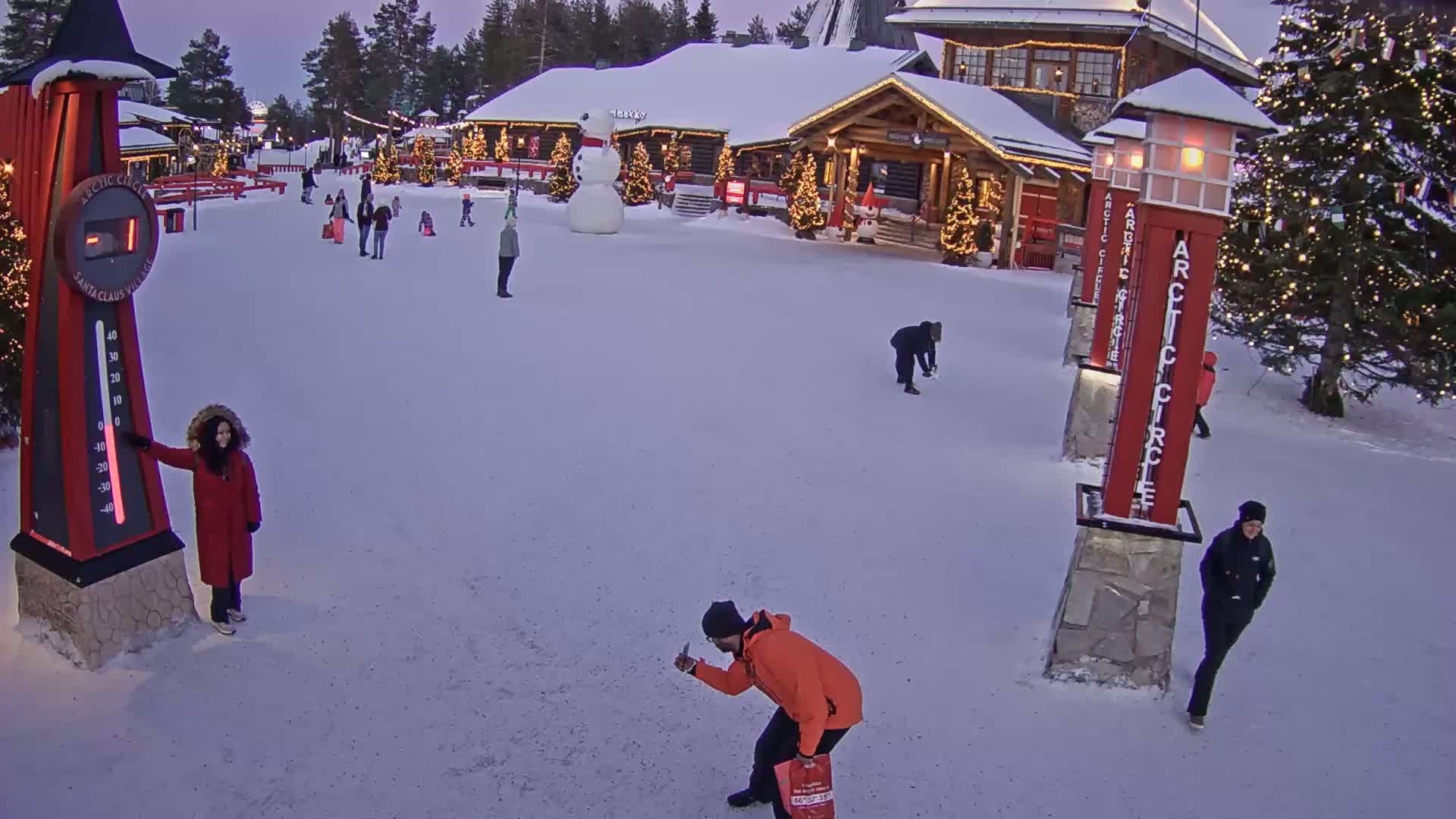 A snow-covered village scene at twilight features several people walking around buildings decorated with Christmas lights, a large snowman, and a tall thermometer indicating a sub-zero temperature.