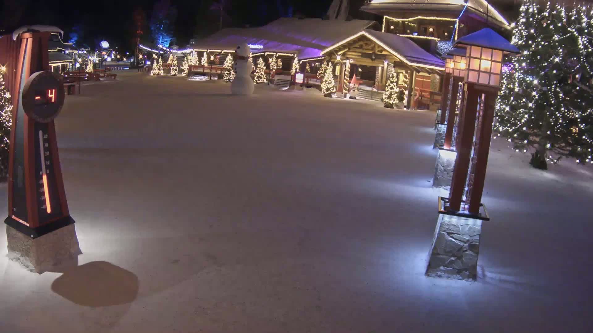 A snow-covered nighttime scene shows a wooden building decorated with Christmas lights, a snowman, and illuminated lampposts at -4 degrees.