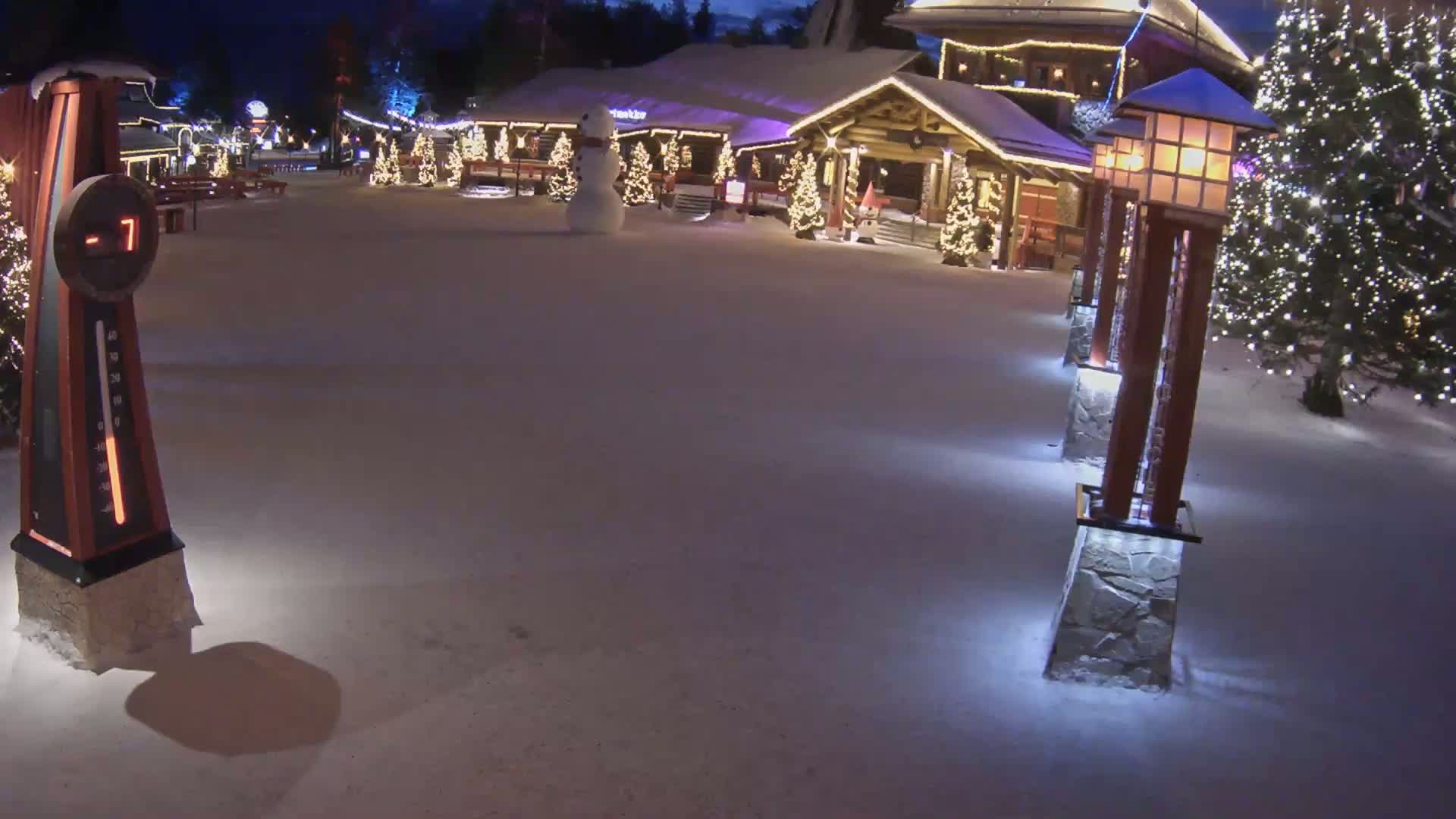 A snow-covered plaza at night, illuminated by lights on posts and Christmas trees, features a snowman in front of a festively decorated building, with a thermometer showing a temperature of -7 degrees.
