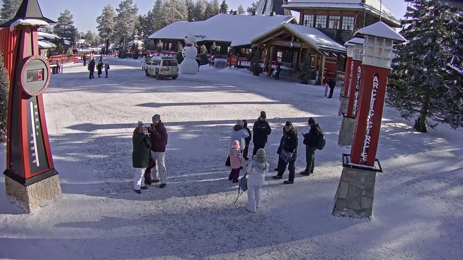 A snowy plaza with several people standing in groups, a large snowman, and buildings in the background under a sunny sky.