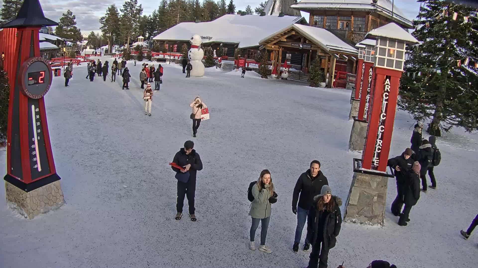 A snow-covered plaza features several people walking around buildings, a large snowman, and a thermometer showing -2 degrees.