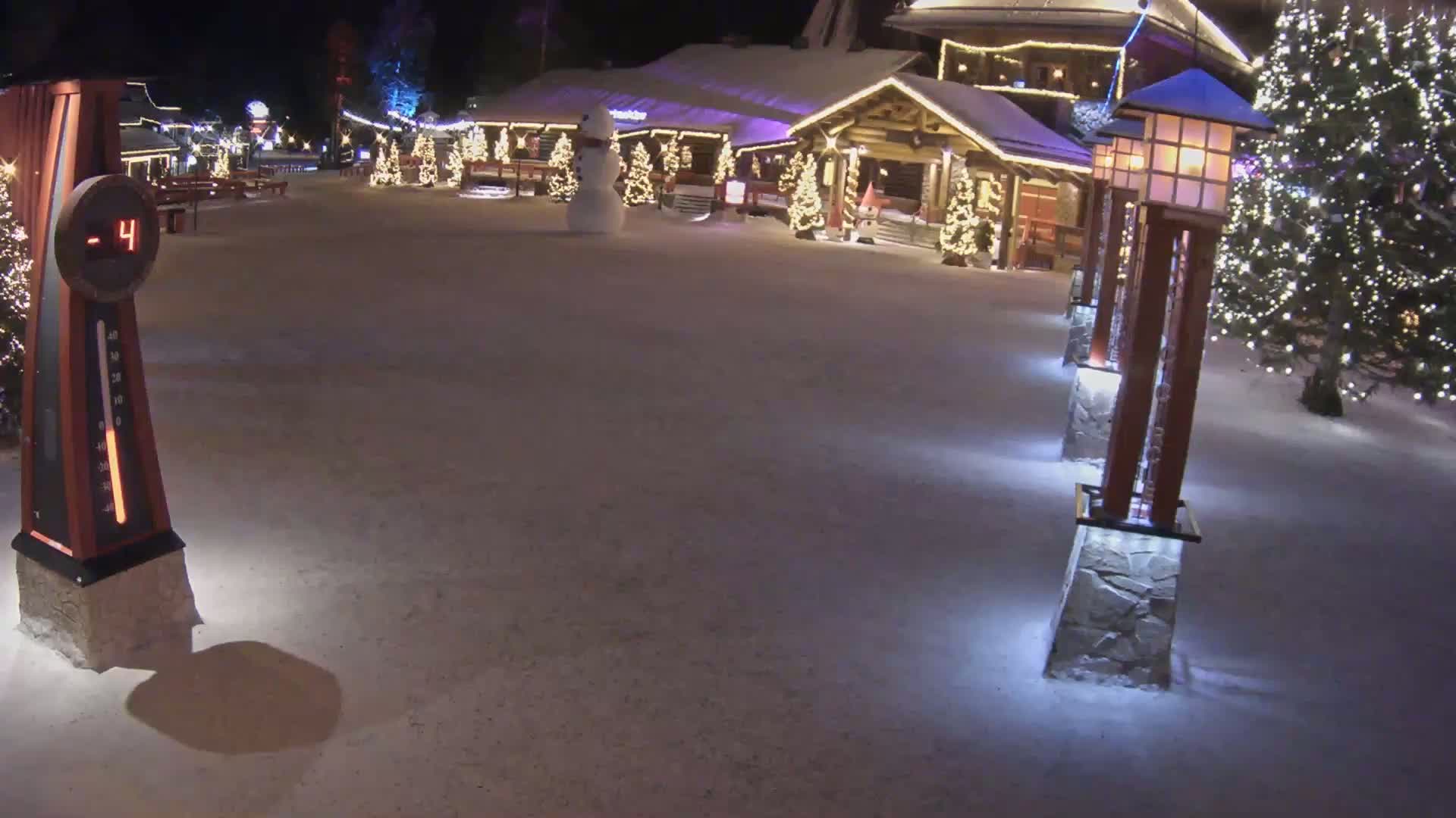 A snow-covered plaza at night, illuminated by lights on buildings and lampposts, features a snowman and a thermometer showing -4 degrees.
