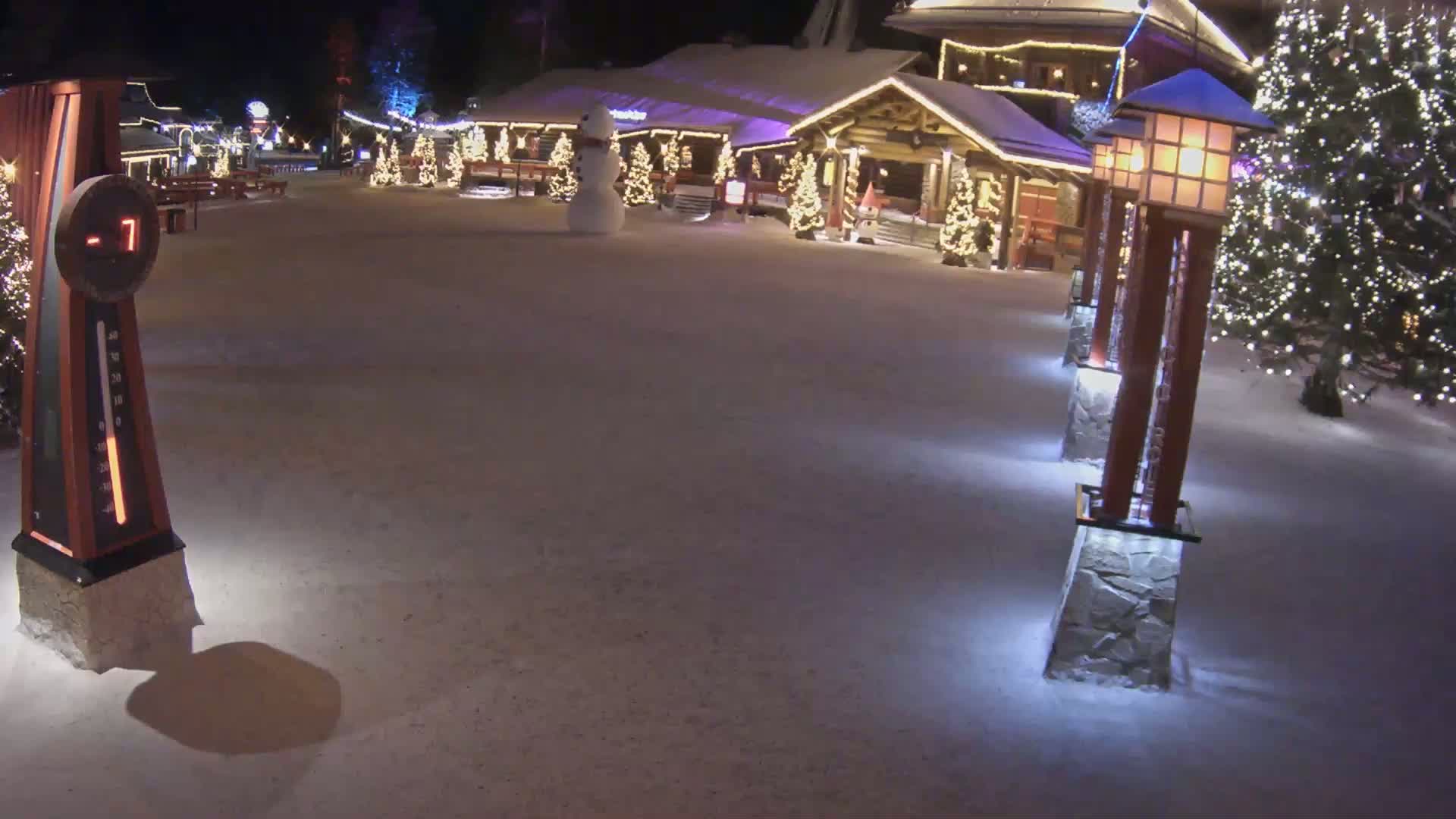 A snow-covered plaza at night is illuminated by lampposts and Christmas lights, featuring a snowman and a building in the background.