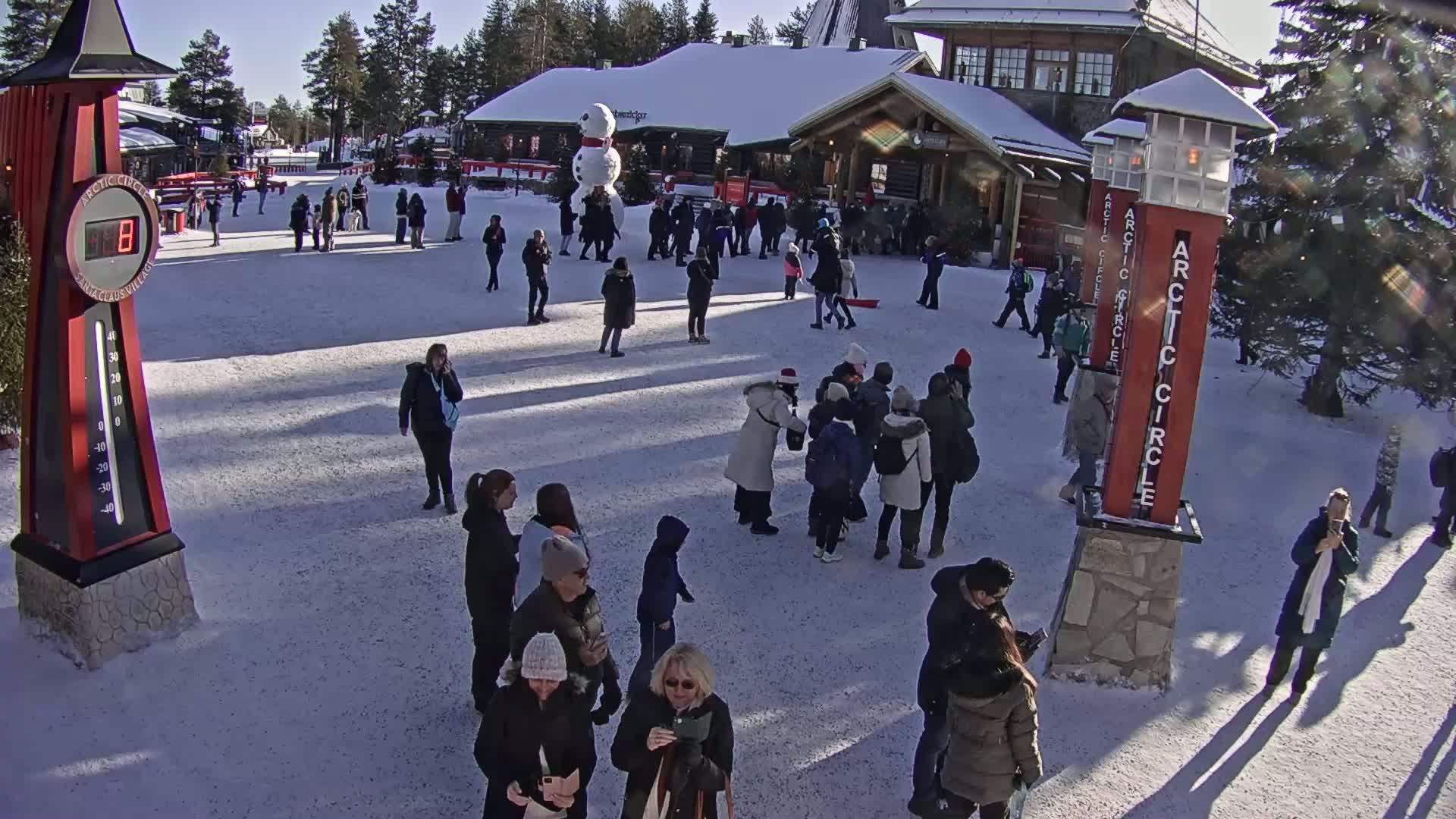 A snowy plaza with many people walking around near wooden buildings under a sunny sky.