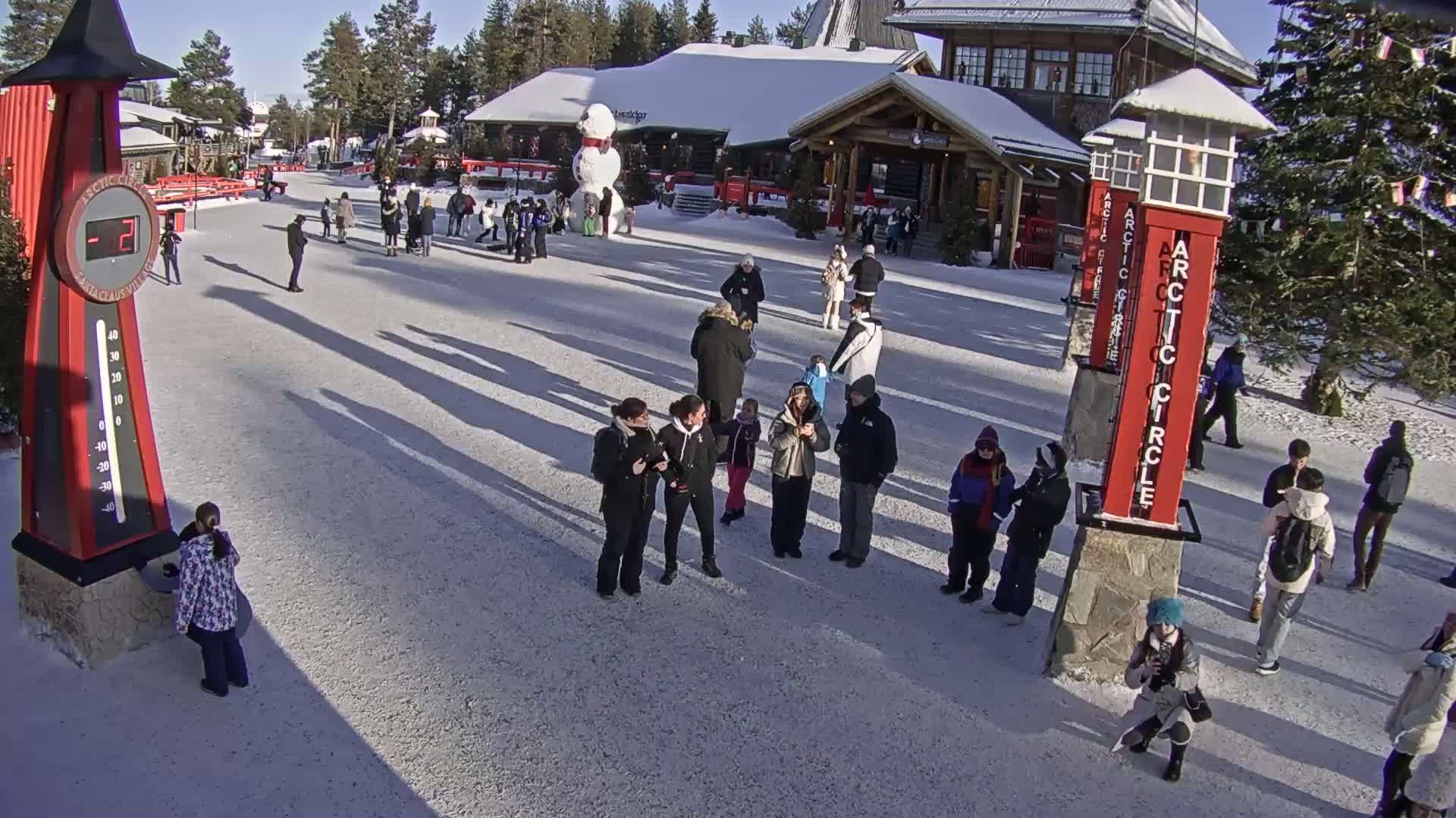 A snowy plaza, filled with people, features a large snowman and several tall red structures, all under a bright, sunny sky.