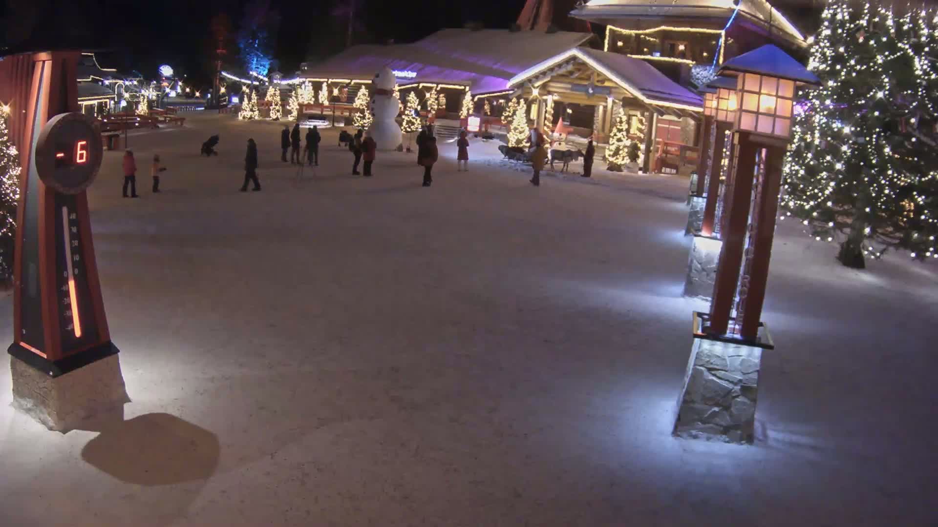 A snow-covered plaza at night is illuminated by Christmas lights, featuring several people walking near festively decorated buildings and a large snowman.