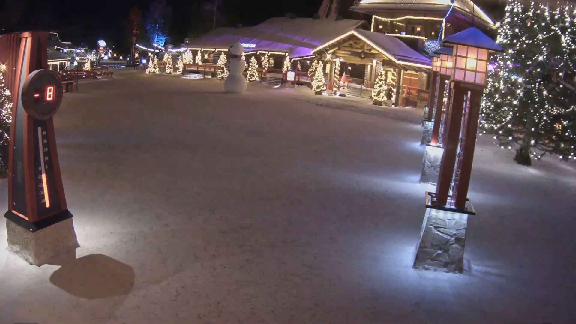 A snow-covered plaza at night features illuminated Christmas trees, decorative lights on buildings, a snowman, and several lighted lampposts, with a thermometer displaying a temperature of -8 degrees.