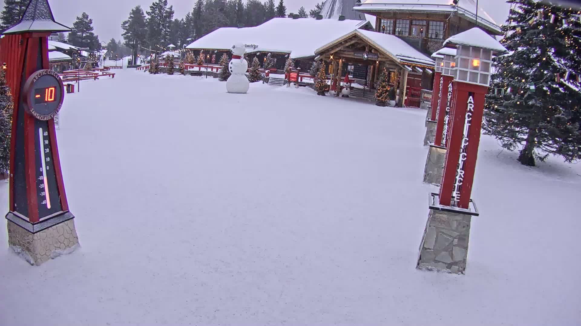 A snow-covered village scene shows a building with Christmas decorations, a snowman, and several tall red markers displaying "Arctic Circle" in the overcast weather.