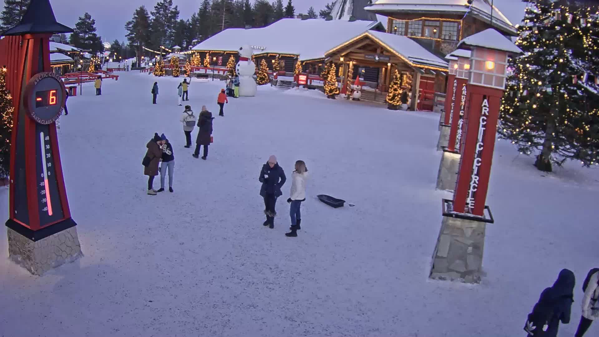 A snow-covered village square with several people walking around, a large snowman, and Christmas trees, under a twilight sky, shows a temperature of -6 degrees.