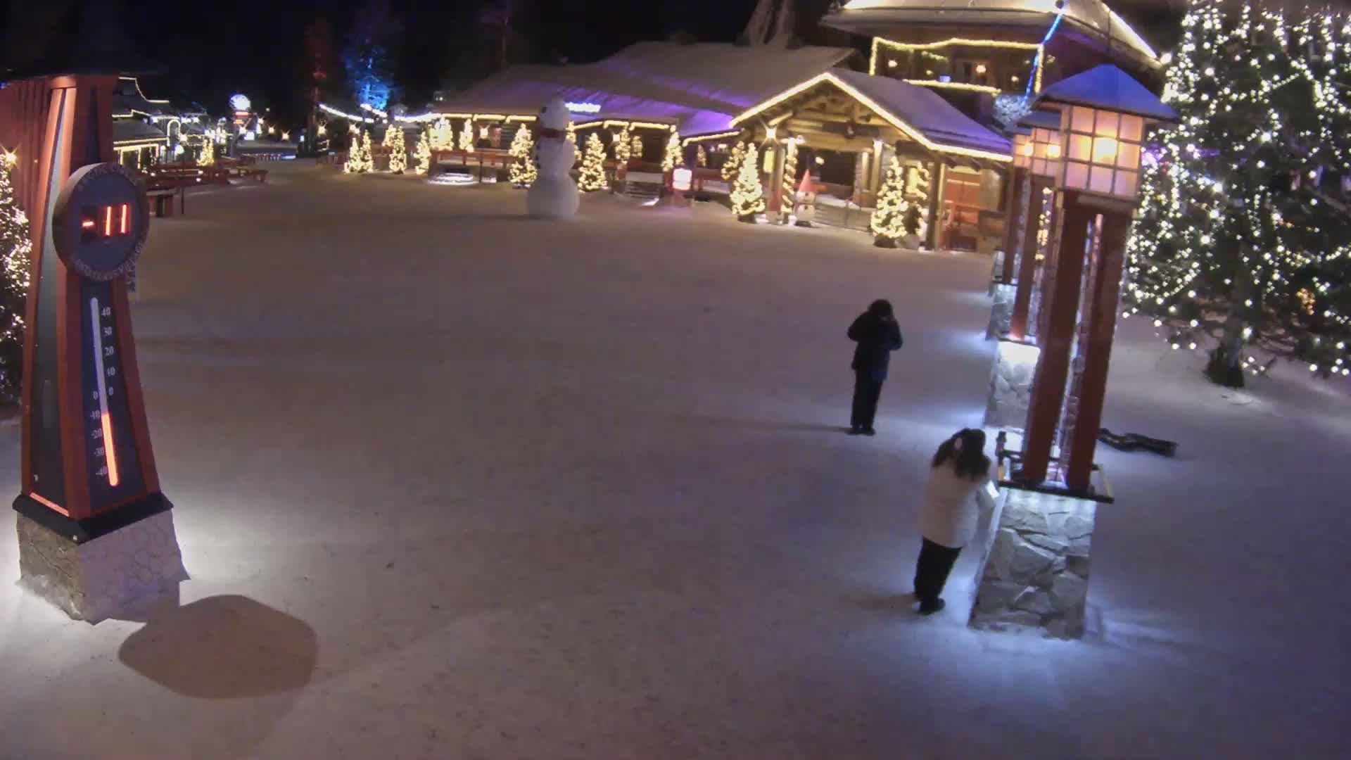 A snow-covered plaza at night is illuminated by Christmas lights, featuring several people standing near tall light posts and a snowman in front of a building.