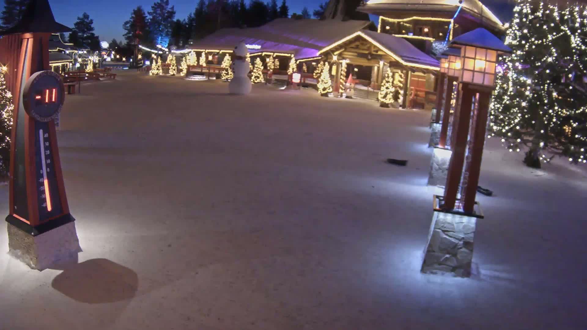 A snow-covered village square at night is illuminated by lights on buildings and lampposts, with a thermometer showing -11 degrees and a snowman in the foreground.