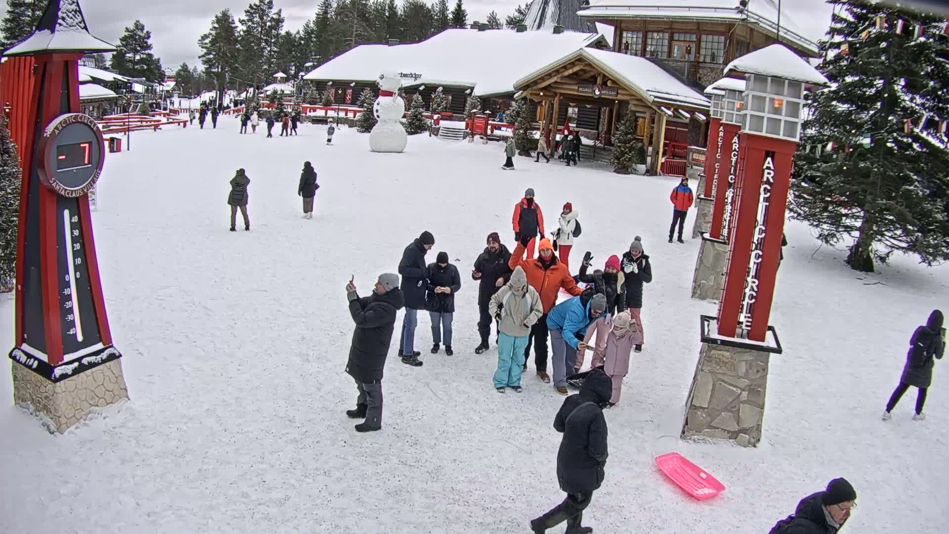 A snowy plaza scene shows several groups of people gathered near red Arctic Circle signposts, a large snowman, and wooden buildings, with a thermometer displaying a temperature of -7 degrees.