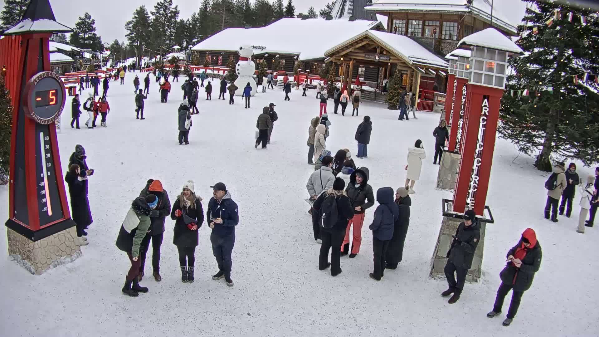 A snowy outdoor plaza with many people milling about near wooden buildings, a large snowman, and tall red Arctic Circle markers; the temperature is -5 degrees.