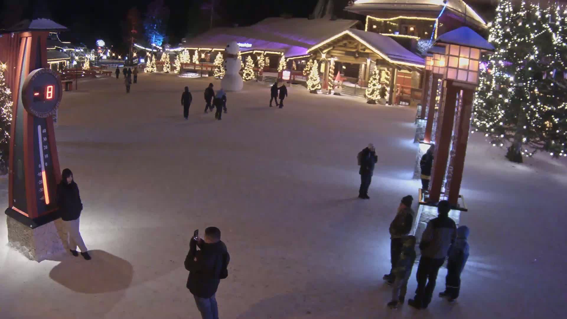 A snow-covered plaza at night, illuminated by lights on buildings and trees, shows several people walking around and a large snowman.