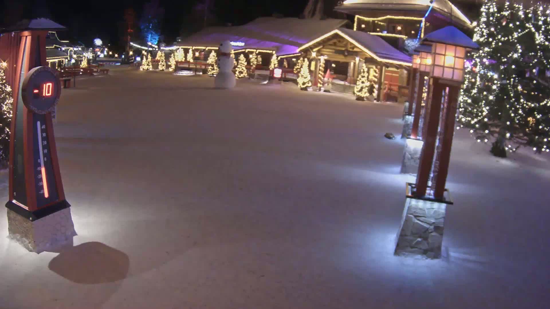 A snow-covered village square at night shows a large thermometer indicating -10 degrees, illuminated light posts, a snowman, and festively lit buildings.