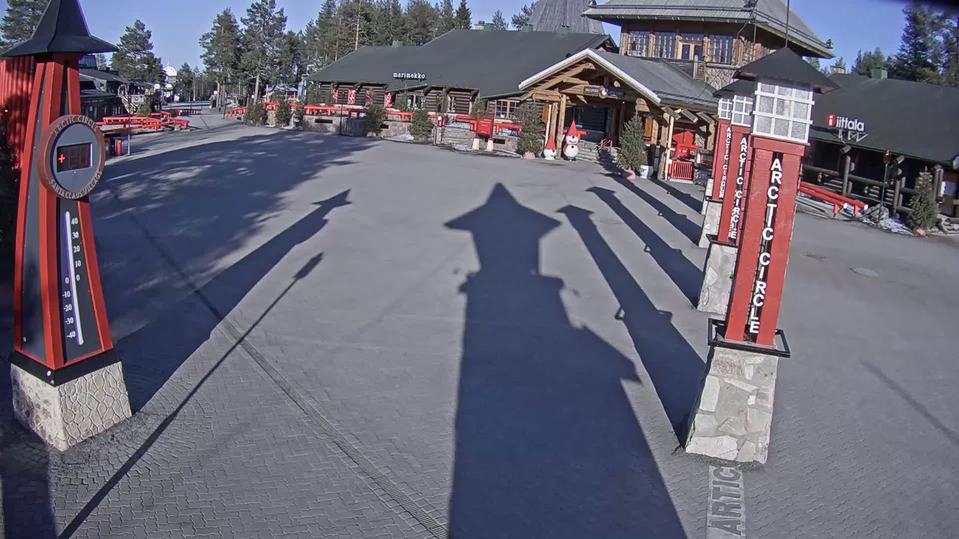 A paved plaza with several tall, red and white posts casting long shadows on a sunny day in front of a log cabin-style building.
