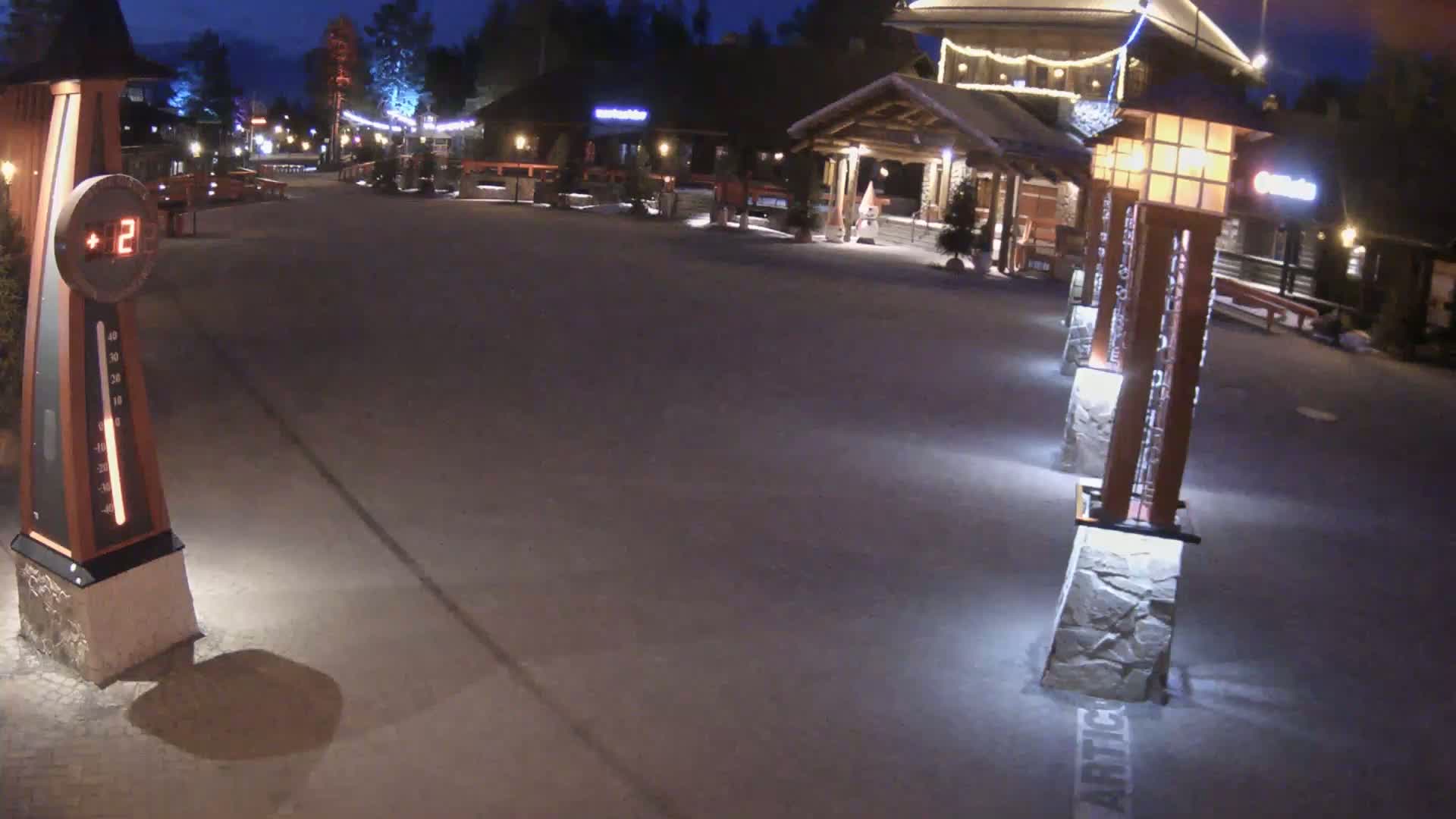 A snow-covered plaza at night, illuminated by lights on tall posts, shows buildings and a thermometer indicating a temperature of -12 degrees.
