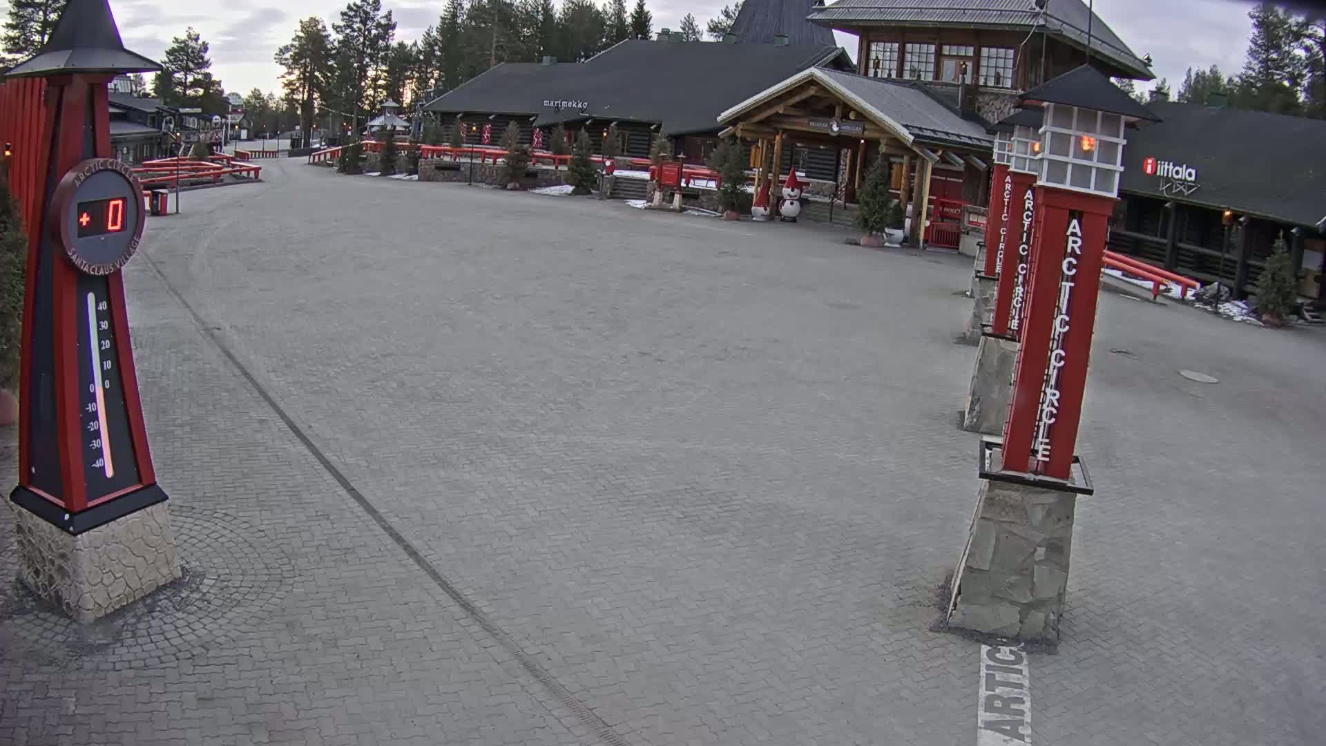 A paved plaza in front of several log buildings under an overcast sky, with a thermometer showing 0 degrees.