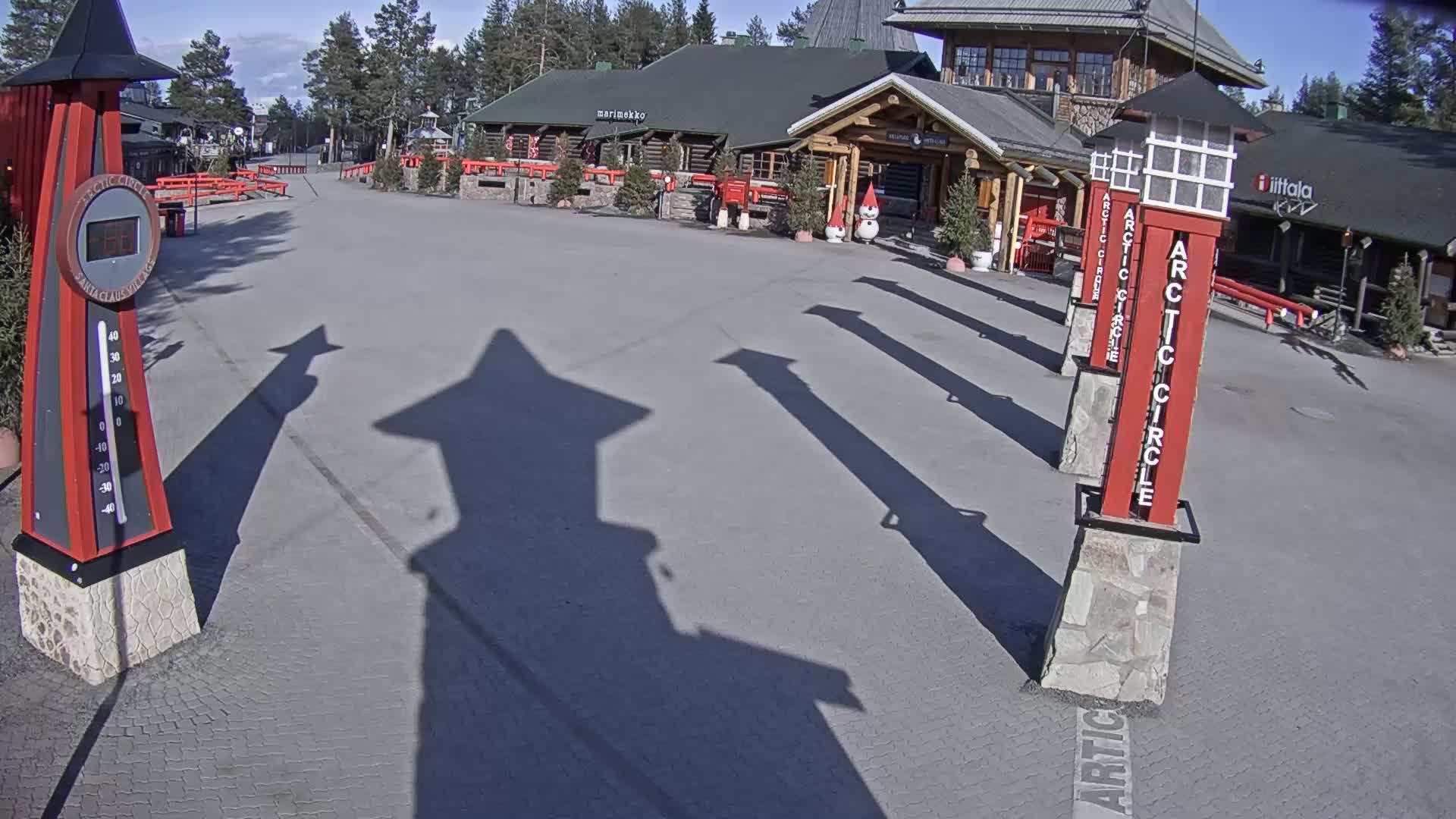 A sunny outdoor scene shows a paved plaza with several tall, red, stone-based posts and the shadows of those posts cast on the ground in front of a low, long log building.