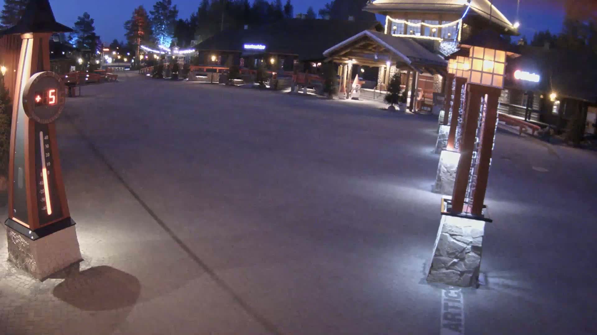 A nighttime view of a village square with wooden buildings and illuminated lampposts, under a clear, dark sky.