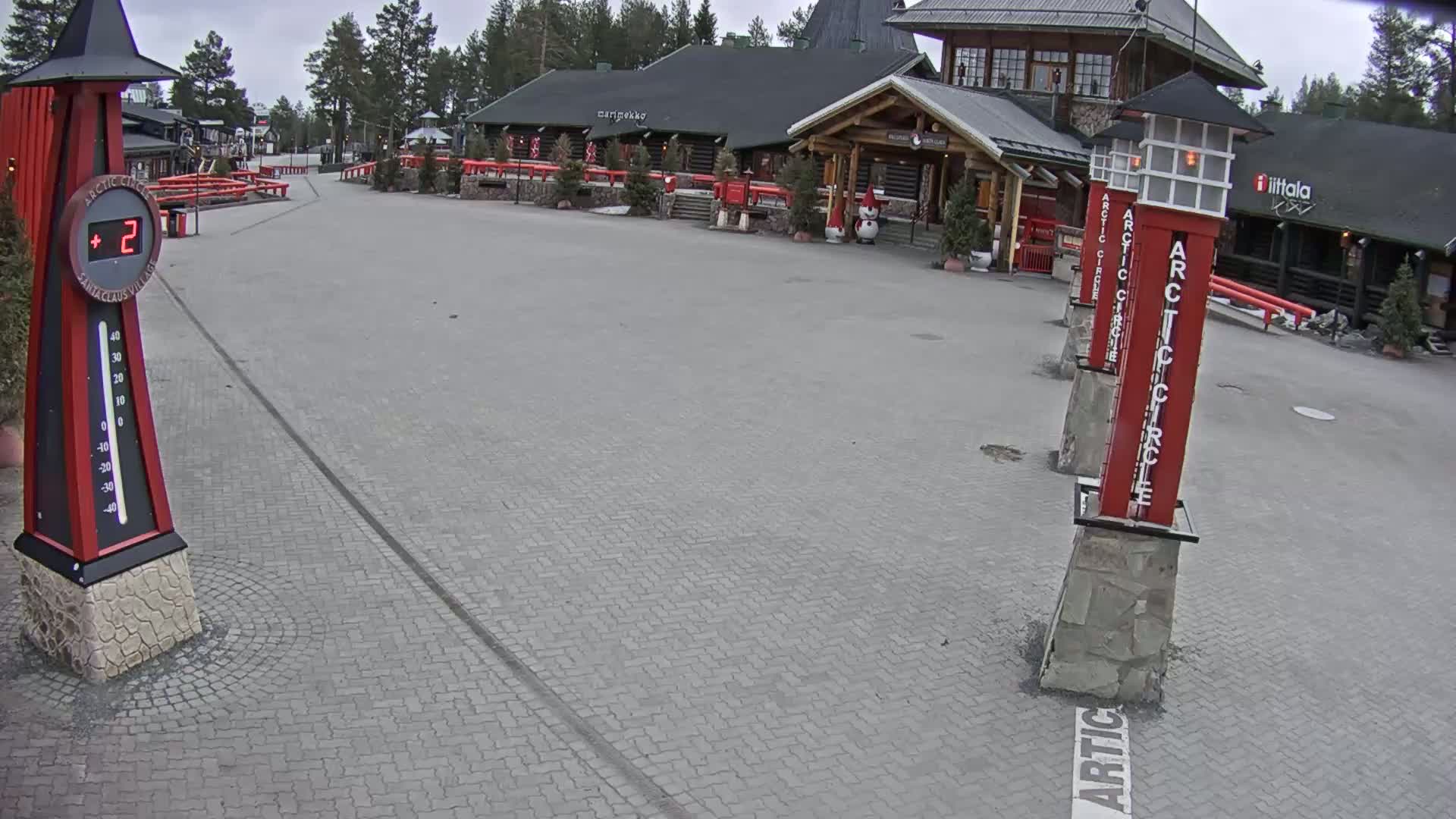 A paved plaza with several red and stone structures, a thermometer showing 2 degrees, and a log cabin-style building in the background on an overcast day.
