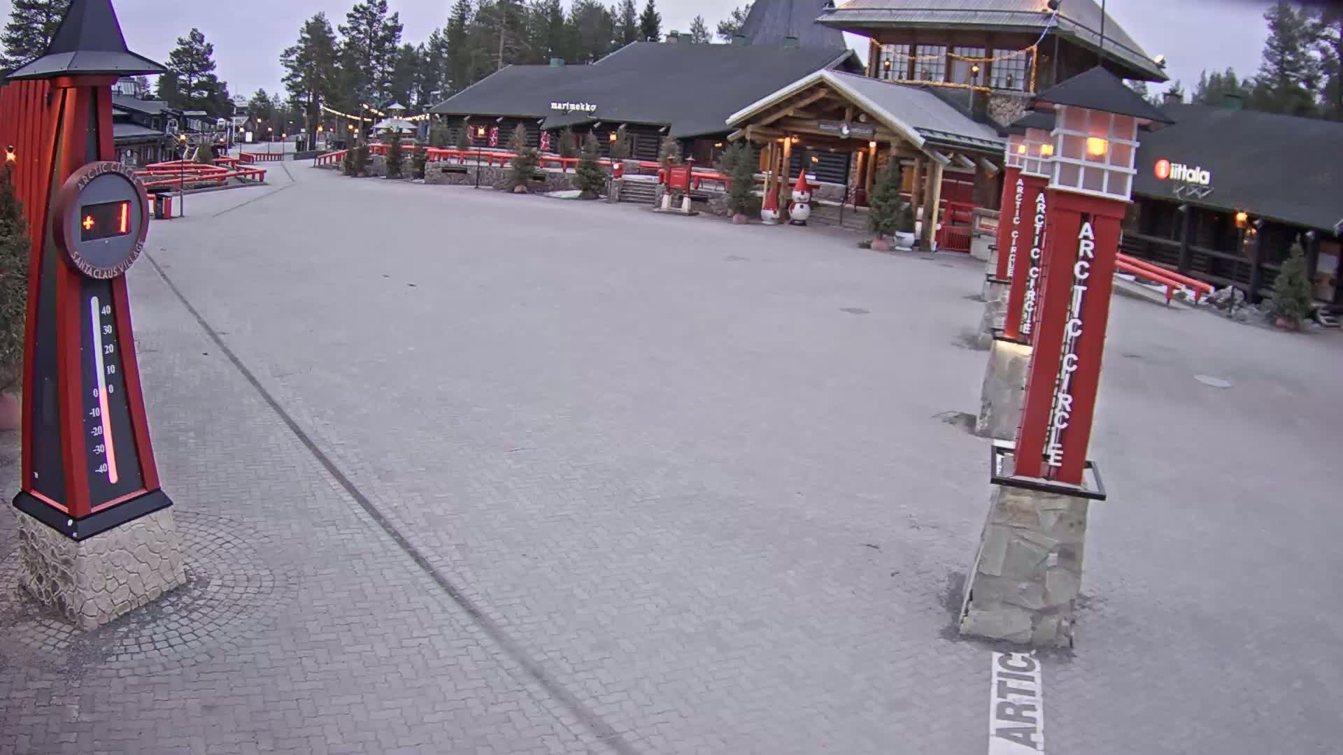 A paved plaza with several log buildings, lit-up light posts, and a large thermometer showing a temperature of one degree, under an overcast sky.