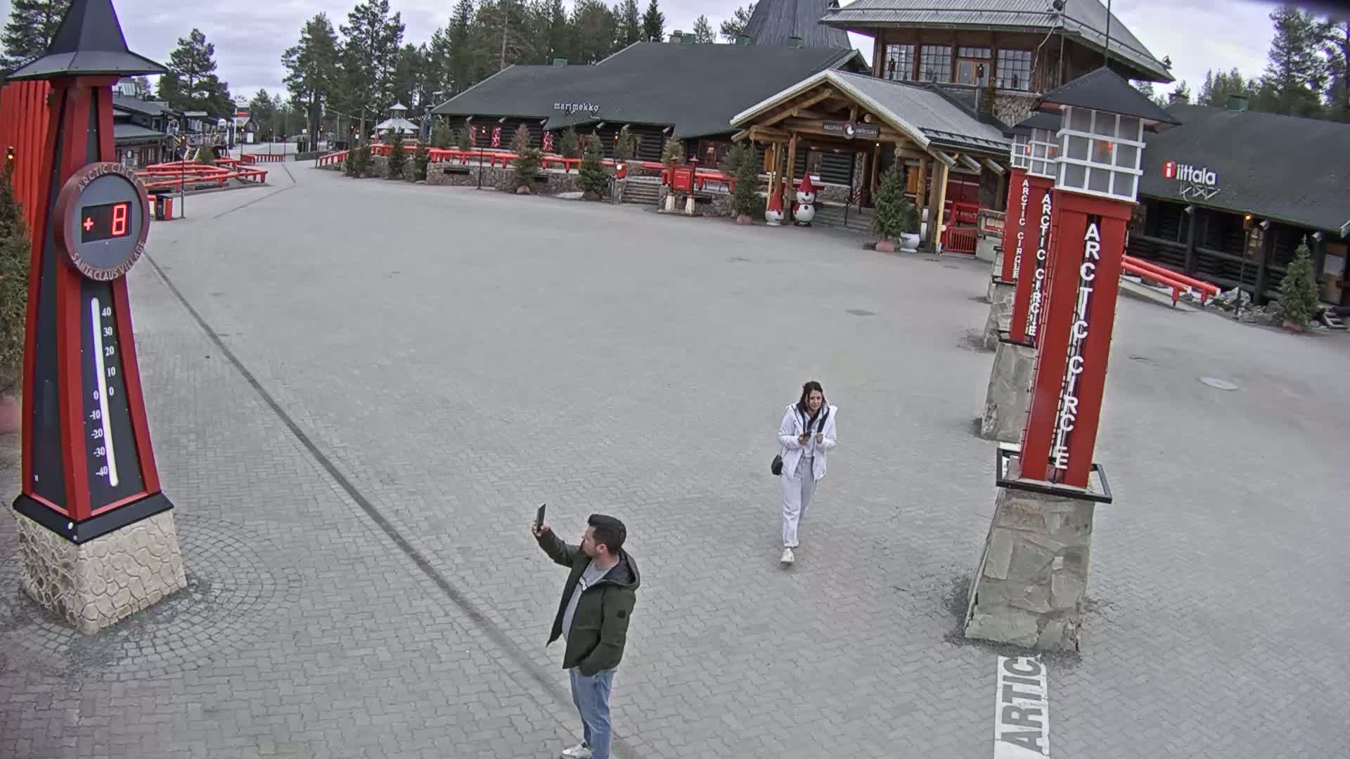 A man takes a selfie while a woman walks past in a paved plaza in front of a log cabin building, under an overcast sky.