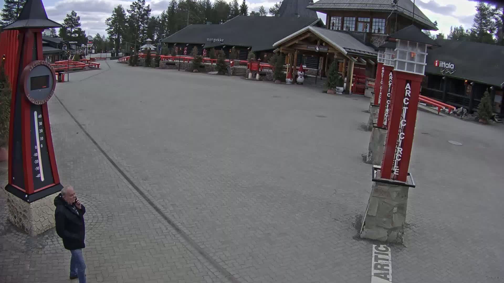 A man stands on a cobblestone plaza in front of a log cabin building under a partly cloudy sky.