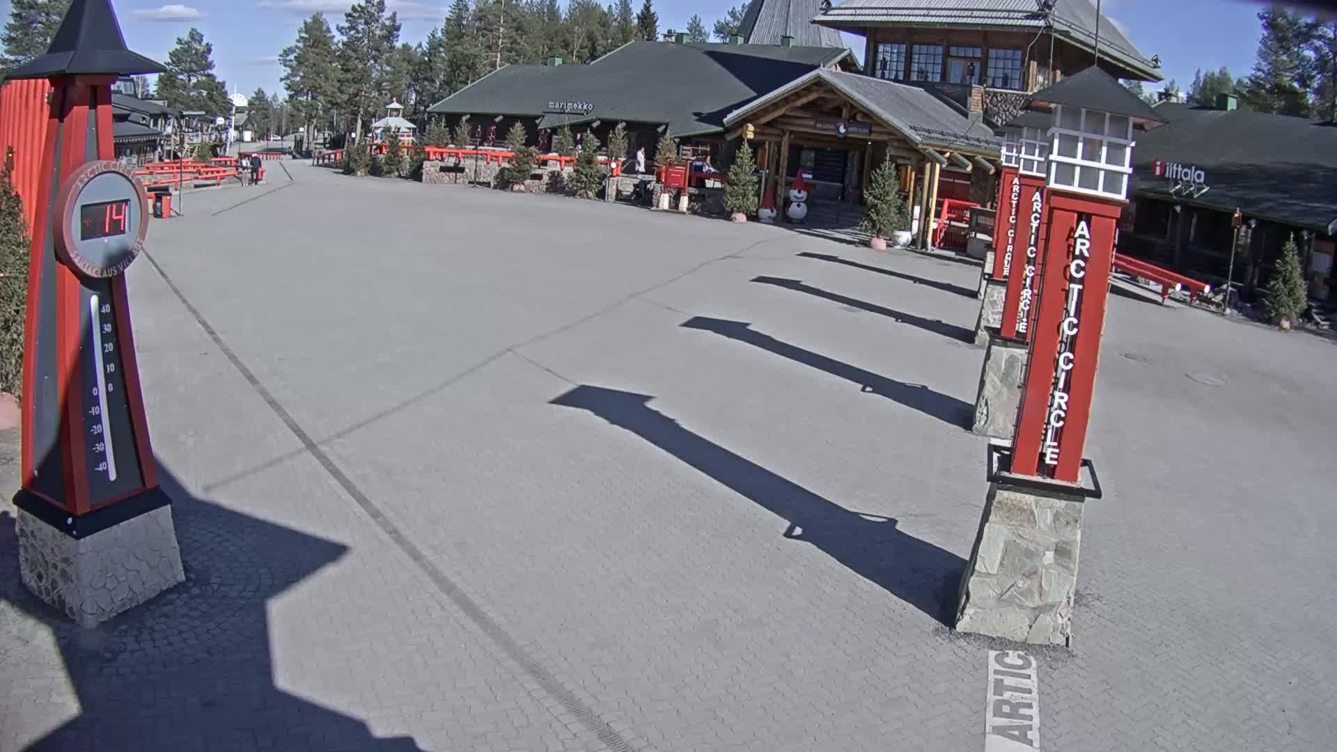 A paved plaza with several tall, red, rectangular pillars, a thermometer showing 14 degrees, and a building in the background on a sunny day.