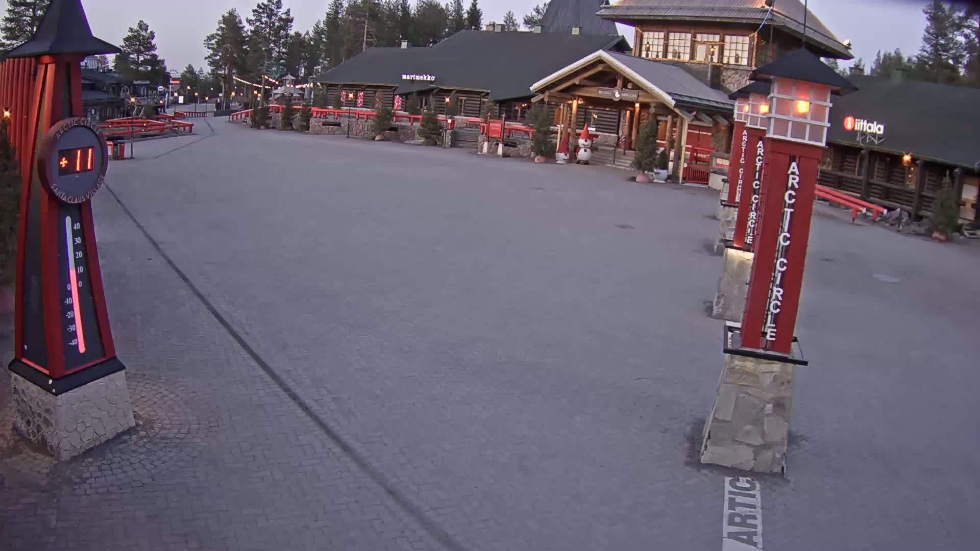 A paved plaza at dusk, showing log buildings and tall, red, rectangular markers with “Arctic Circle” written on them.