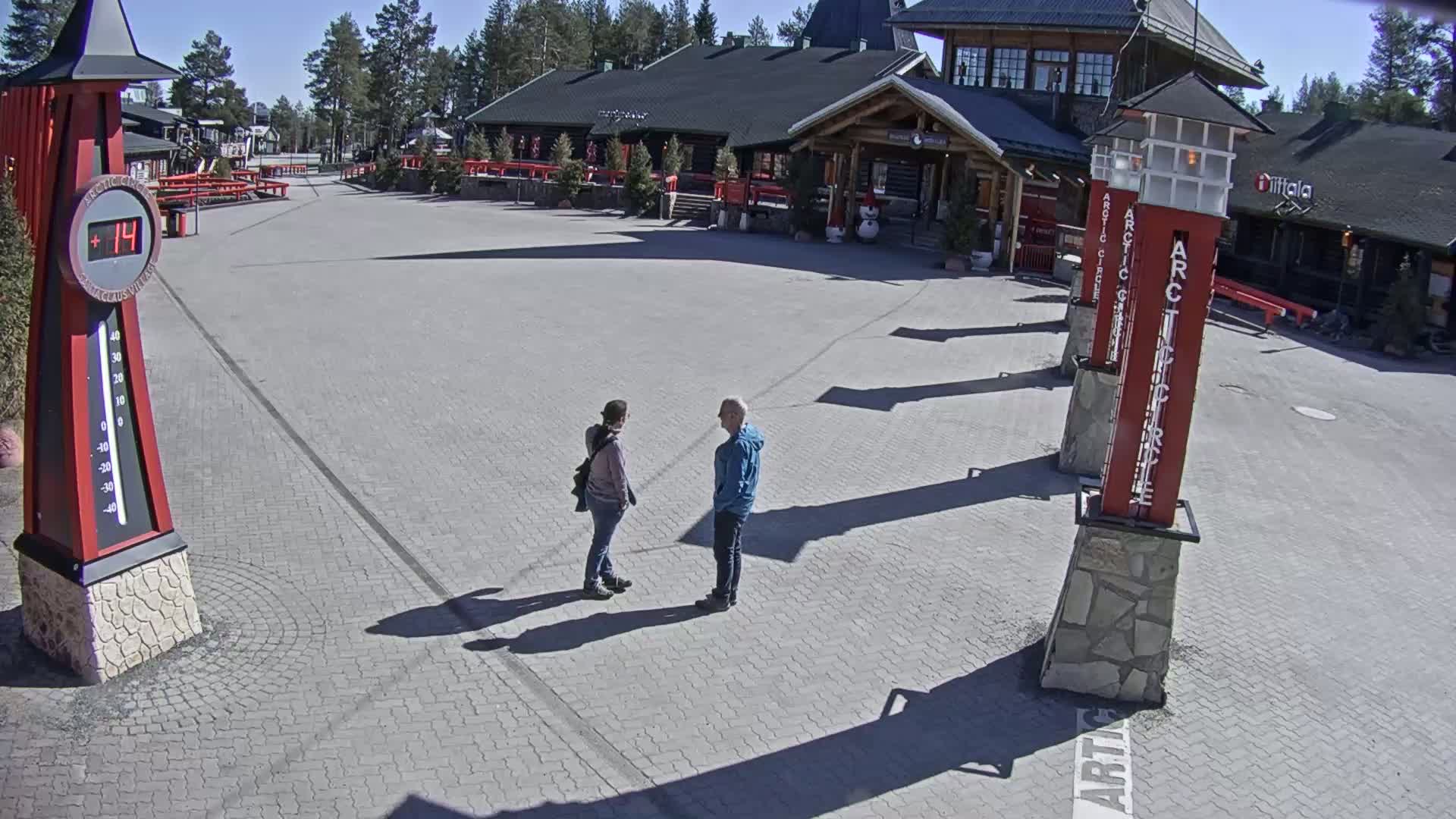 Two people stand in conversation on a sunny day in a paved plaza in front of a log cabin-style building surrounded by trees.