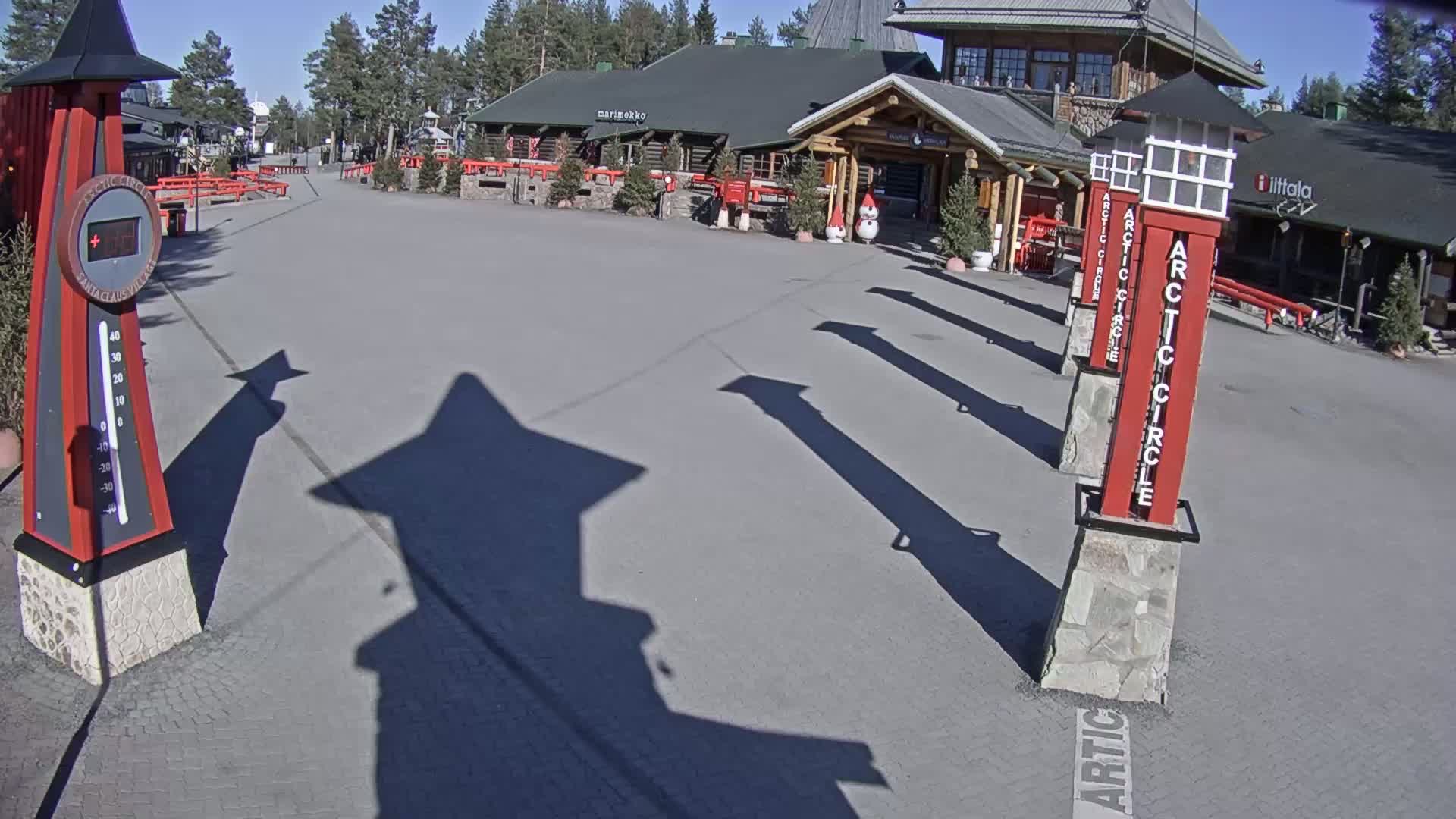 A sunny day reveals a paved area in front of log buildings, with several tall, red and white posts casting long shadows.