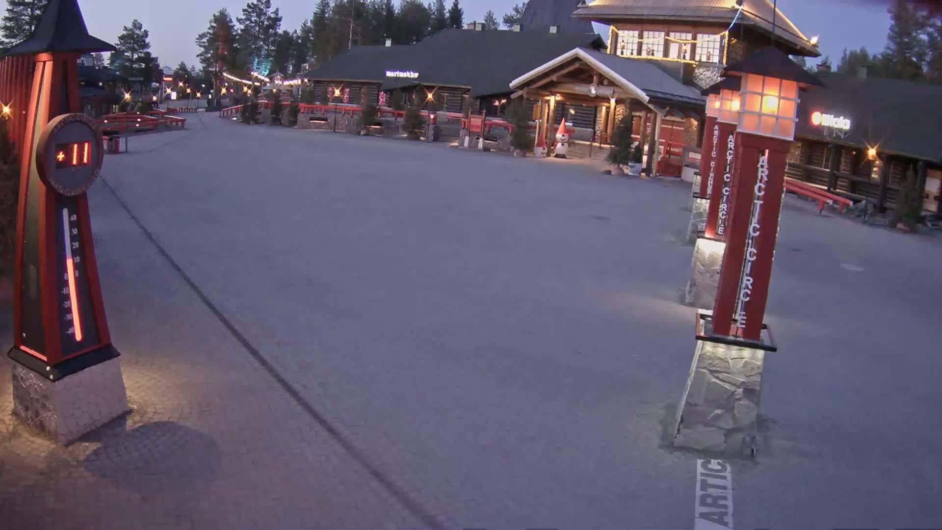 A mostly empty paved plaza at dusk is illuminated by lamps on tall posts, next to a log cabin-style building and a large outdoor thermometer showing 11 degrees.
