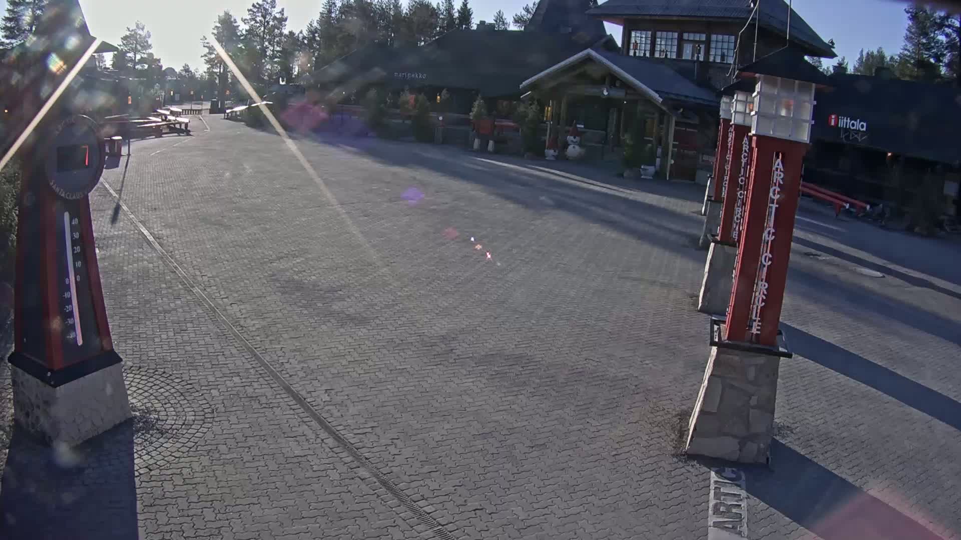 A sunny day reveals a paved plaza in front of a building, with several tall, slender red pillars and a thermometer.