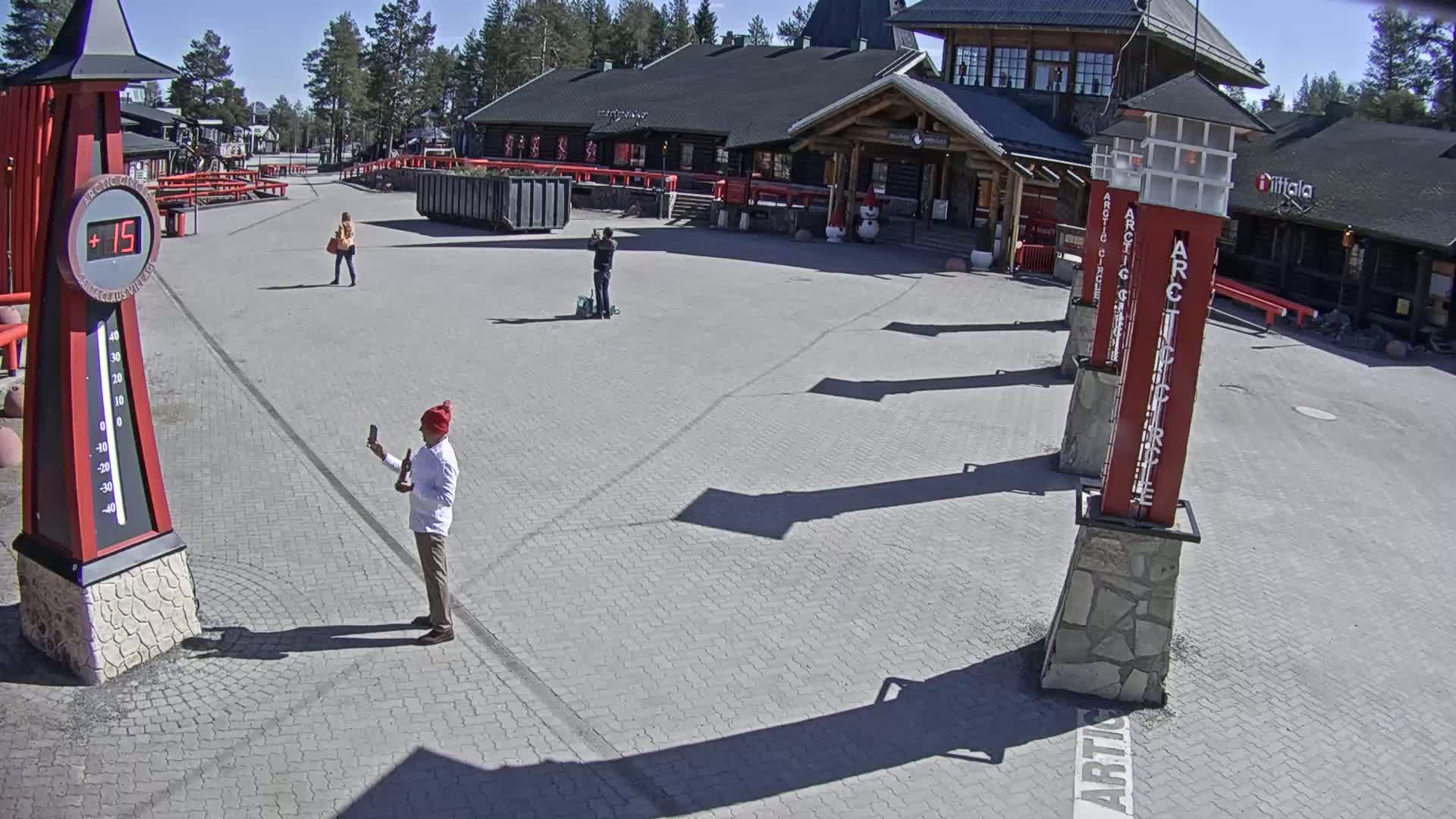 Under sunny skies, a paved plaza features several people, a large thermometer displaying 15 degrees, and tall, slender structures.