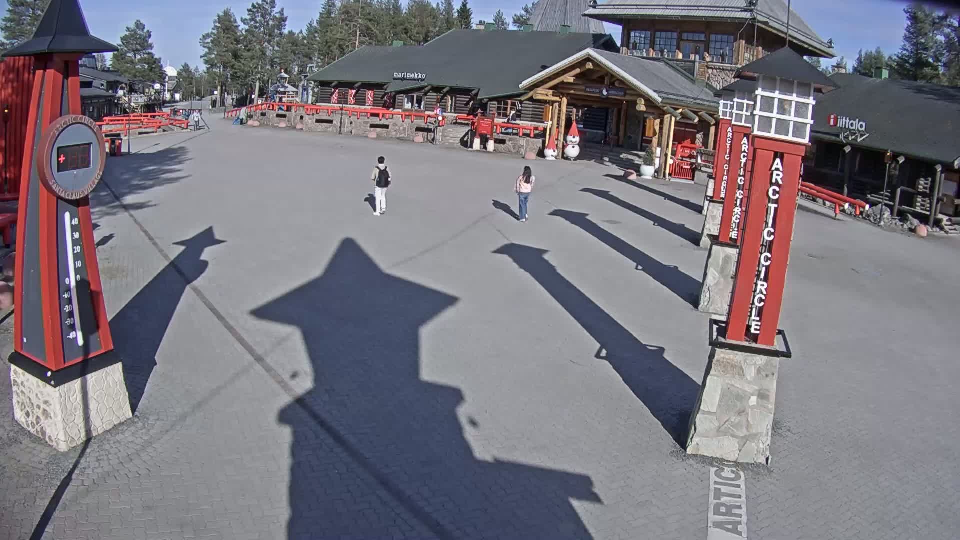 A paved plaza in sunny weather features two pedestrians, several tall red posts with "Arctic Circle" written on them, a thermometer, and a log cabin-style building in the background.