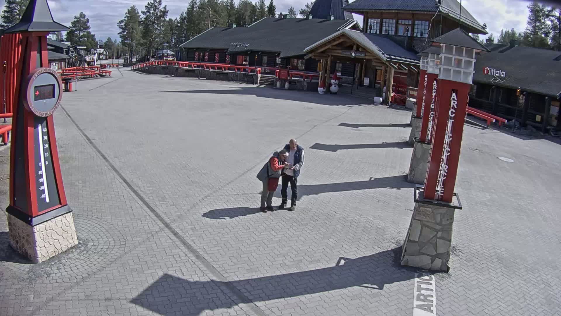 Two people are looking at something on a paved plaza in front of a building on a sunny day, with tall red pillars nearby and a thermometer showing the temperature.