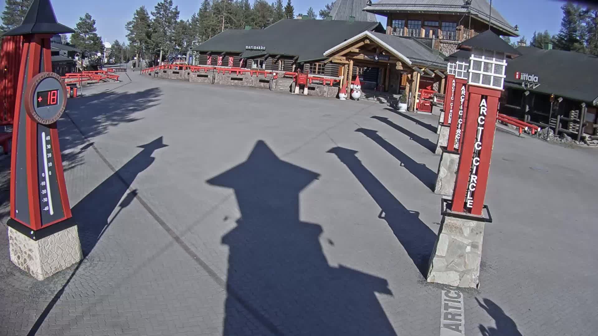 A paved plaza with several red and white Arctic Circle markers and the shadow of one cast on the ground, under a sunny sky, in front of a building.