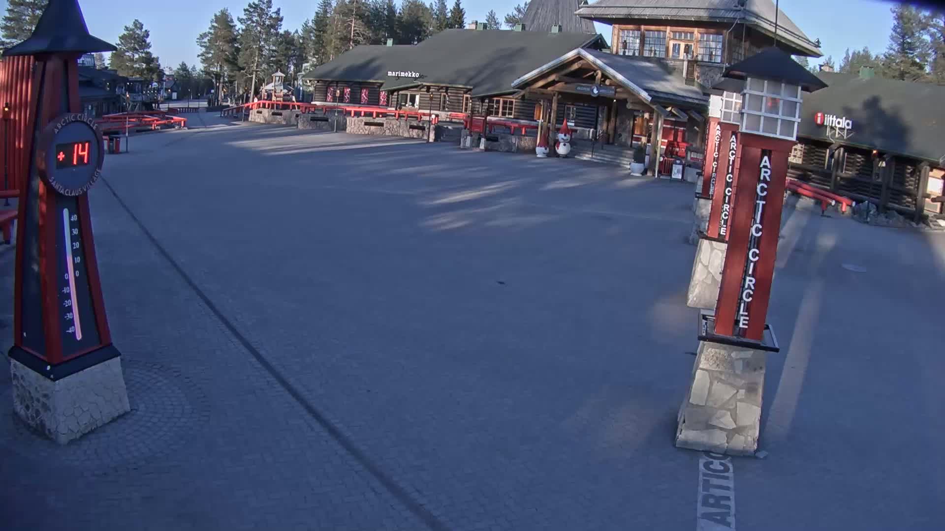 A paved plaza in front of several log buildings under a clear, sunny sky shows a temperature of 14 degrees, with several tall, thin, red signs indicating the Arctic Circle.