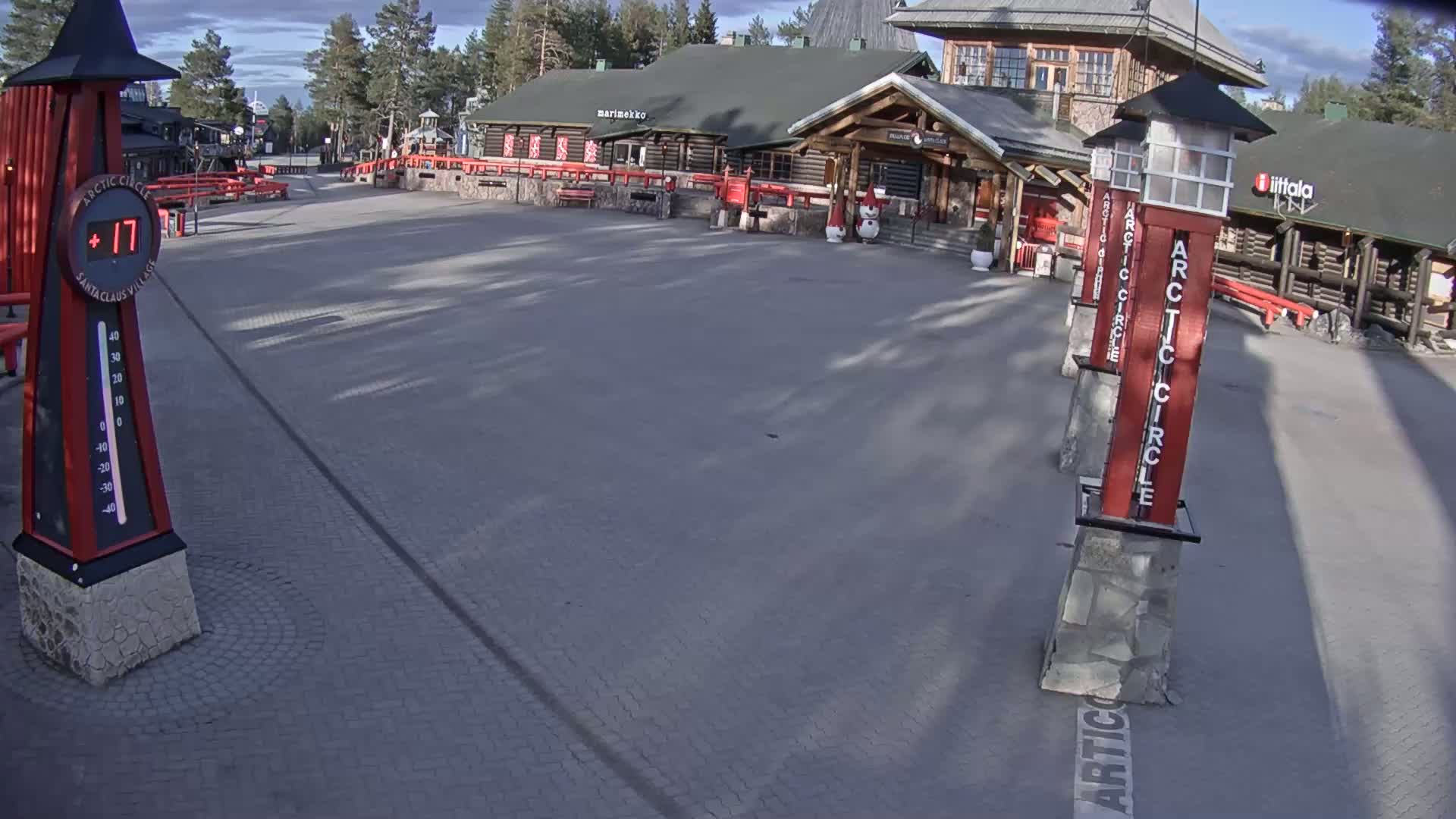 A paved plaza in front of a log building, under a partly cloudy sky, shows a thermometer displaying a temperature of 17 degrees and several tall markers indicating the Arctic Circle.