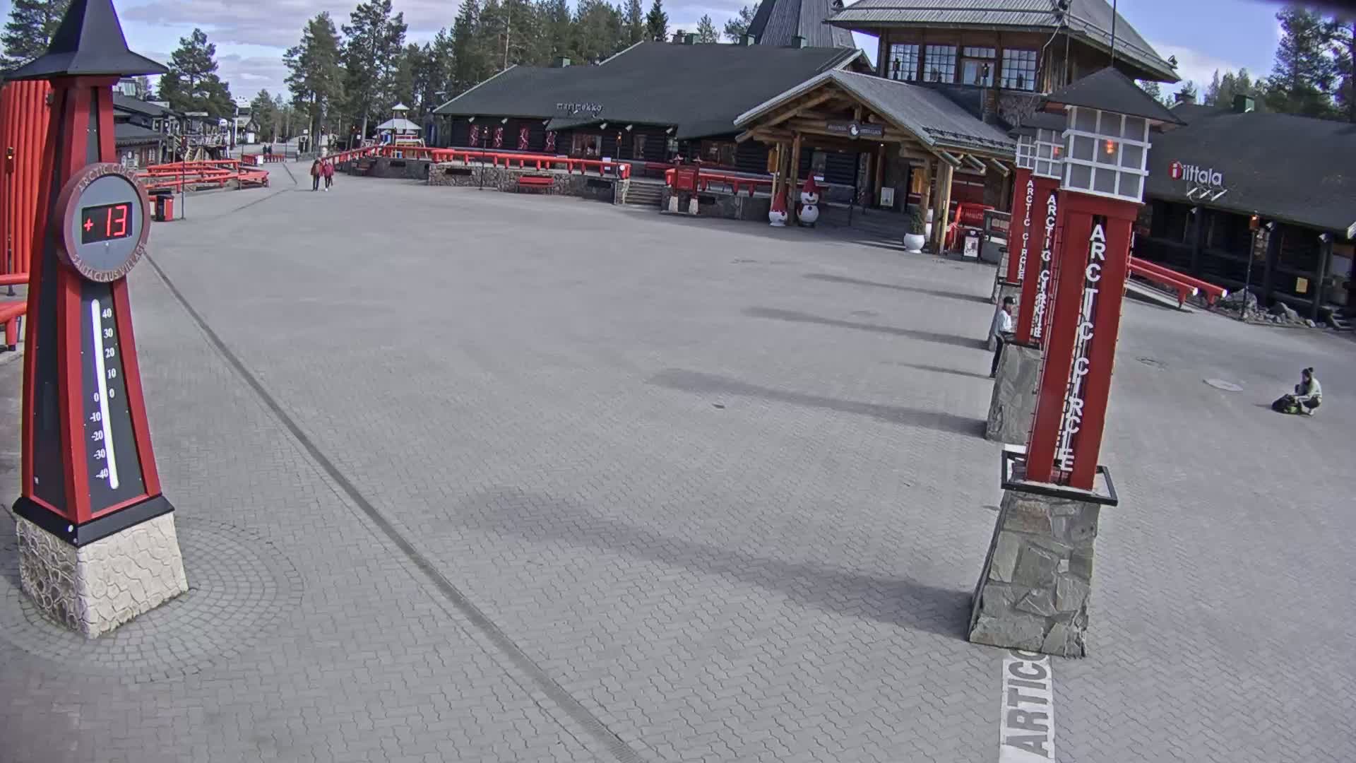 A paved plaza with several buildings, a thermometer showing 13 degrees, and several people is visible under a sunny sky.