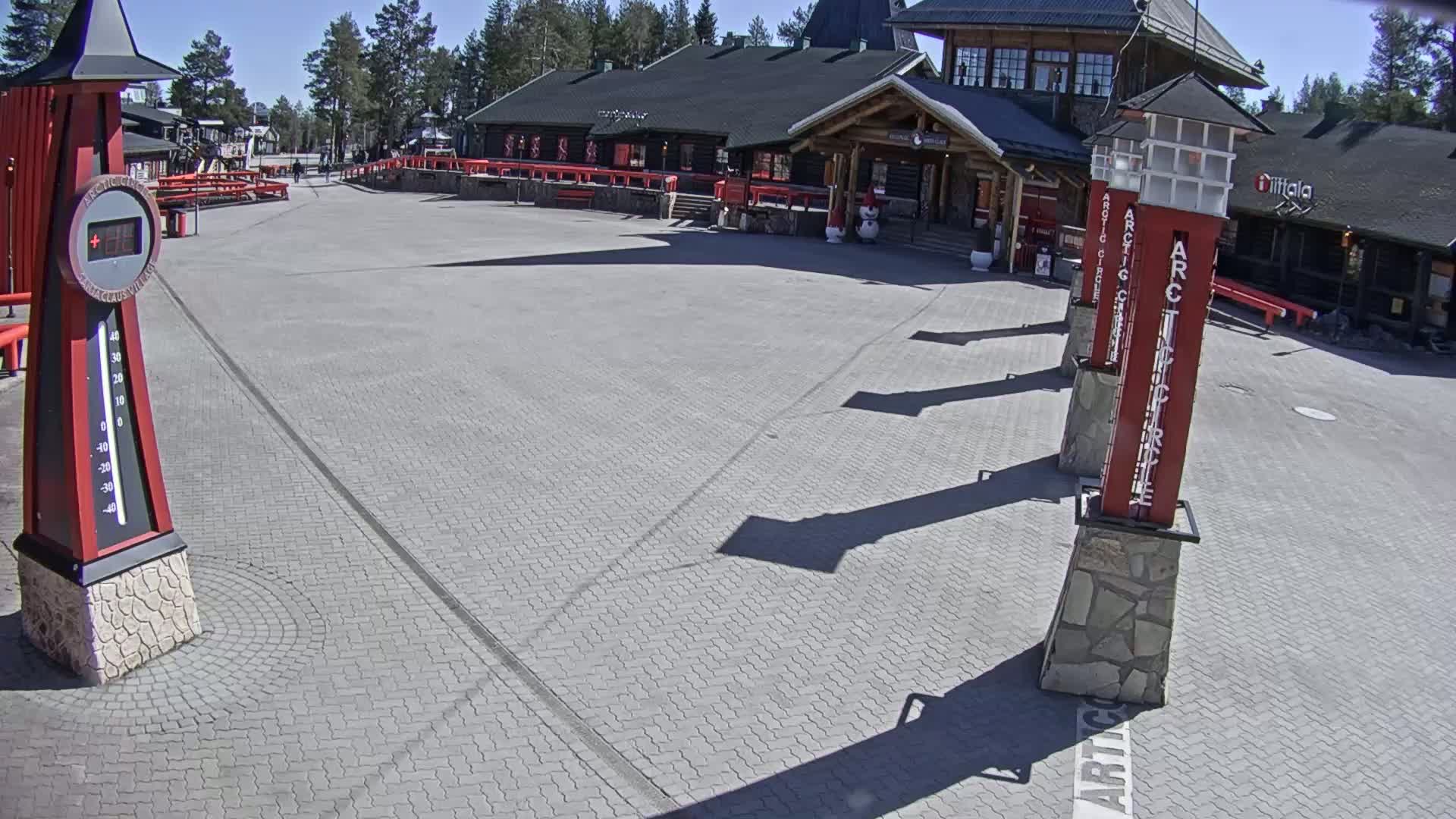 A paved plaza in front of a building on a sunny day, featuring a large thermometer and several tall, slender pillars.