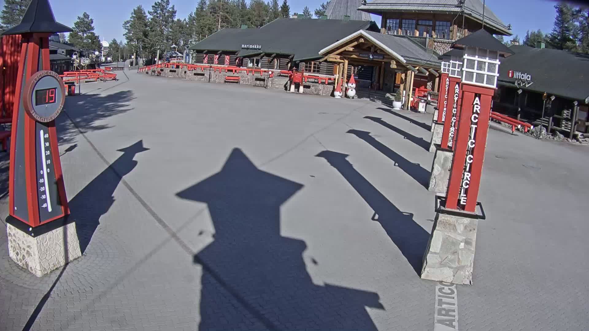 A paved plaza under a sunny sky features a log cabin-style building, several tall red posts marked "Arctic Circle", and a thermometer displaying a temperature of 19 degrees.