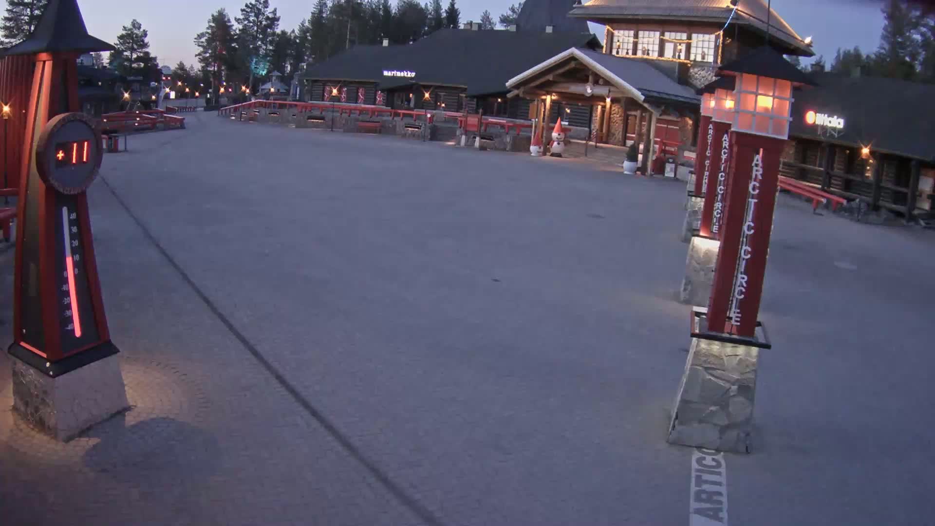 An empty plaza at dusk with several illuminated red and stone pillars, a thermometer showing 11 degrees, and a log cabin-style building in the background.